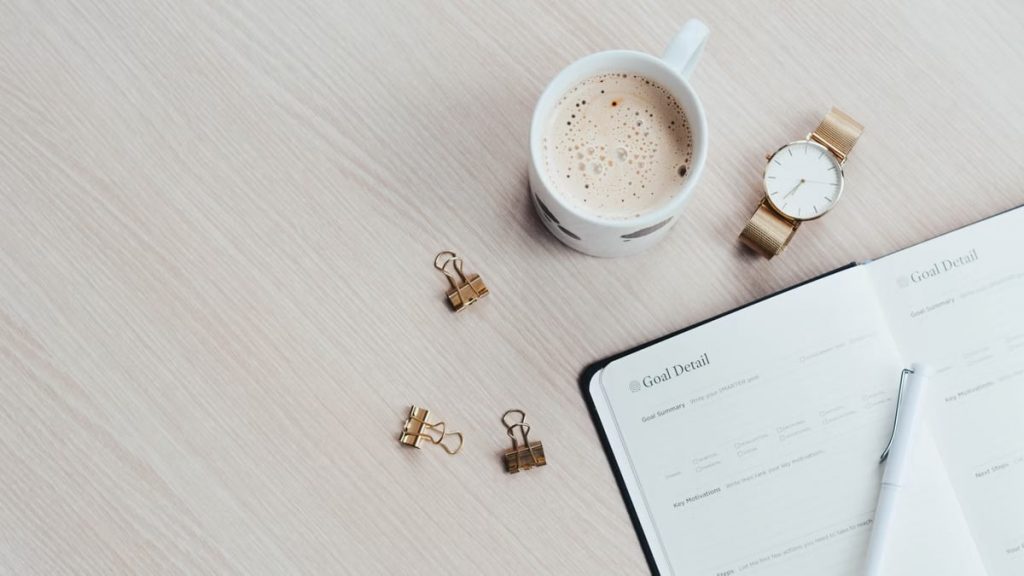 A minimalist desk arrangement featuring a coffee cup, notebook, pen, and wristwatch.