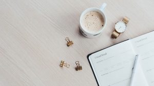 A minimalist desk arrangement featuring a coffee cup, notebook, pen, and wristwatch.