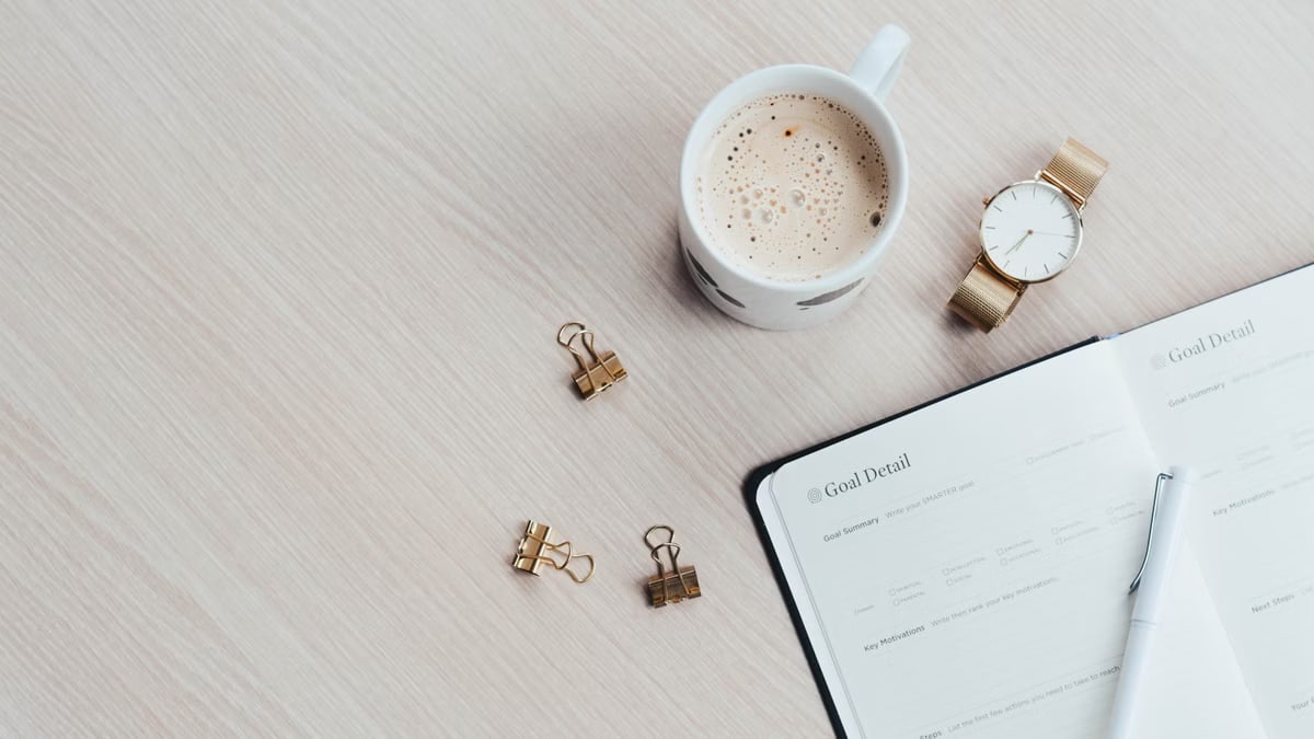 A minimalist desk arrangement featuring a coffee cup, notebook, pen, and wristwatch.